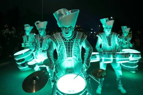 Five performers in shiny, light-up costumes are playing drums at night. A crowd is watching in the background.