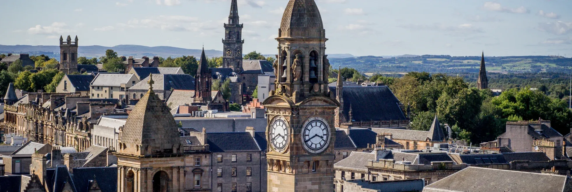 Scenic view of Paisley town centre in Renfrewshire, with the historic clock tower of Paisley Town Hall in the foreground, Victorian and Gothic buildings around it, church spires, and green hills in the distance under a partly cloudy sky.