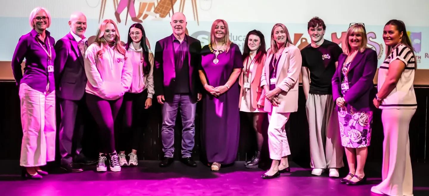 11 people standing and smiling on a purple stage