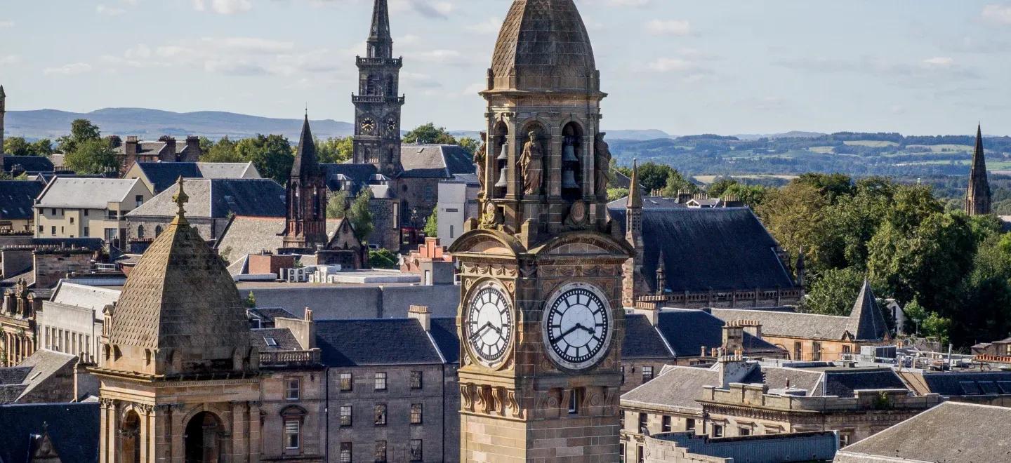 Scenic view of Paisley town centre in Renfrewshire, with the historic clock tower of Paisley Town Hall in the foreground, Victorian and Gothic buildings around it, church spires, and green hills in the distance under a partly cloudy sky.