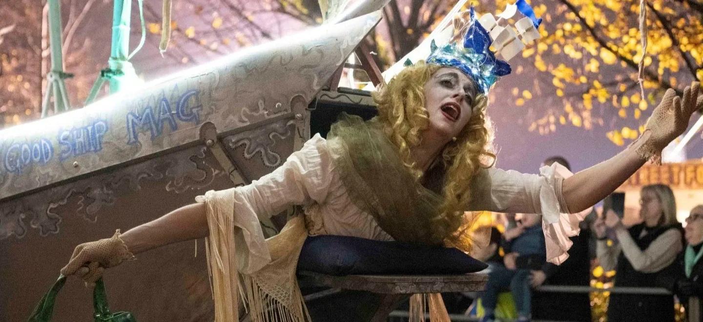 Person in a ship costume labelled "The Good Ship MAG" at a parade, with people watching.