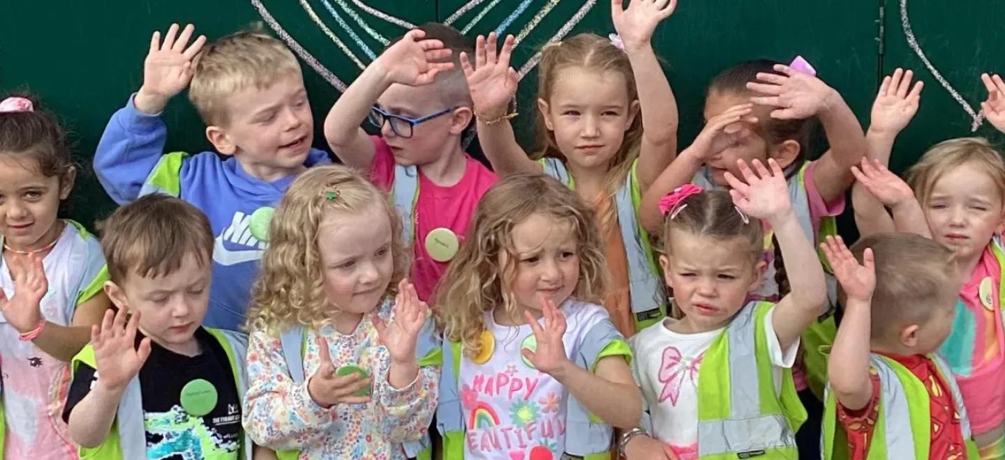 Children from St John Bosco School and ELCC raise their hands in front of a green, heart-decorated backdrop.