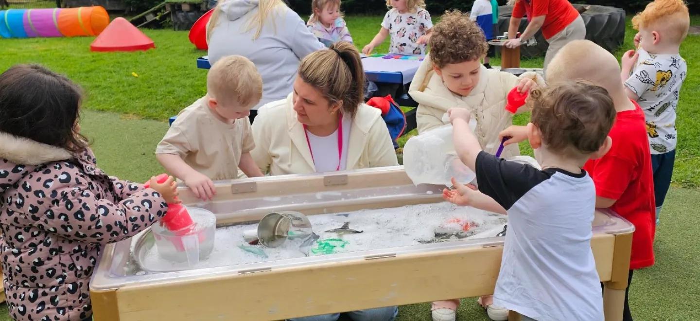 A nursery worker interacts with young children playing in a tub of water.