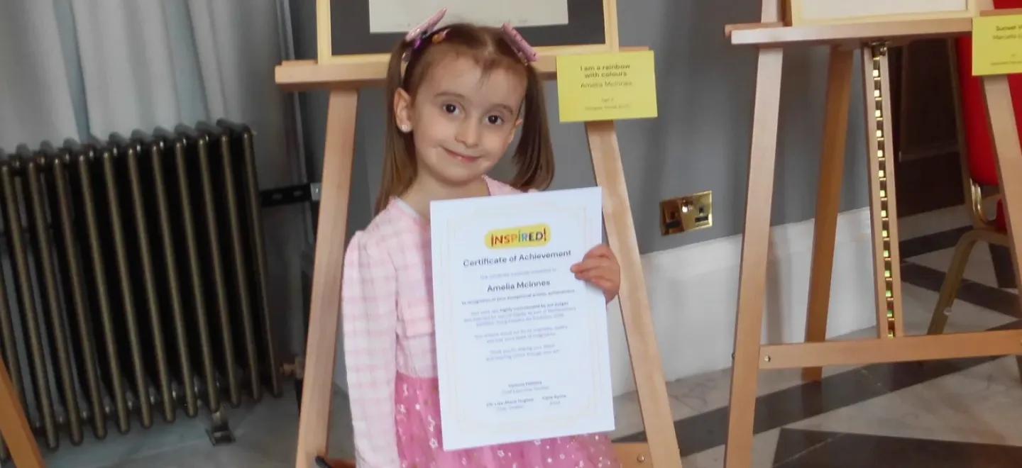 A young child smiles while holding a certificate in an art gallery.
