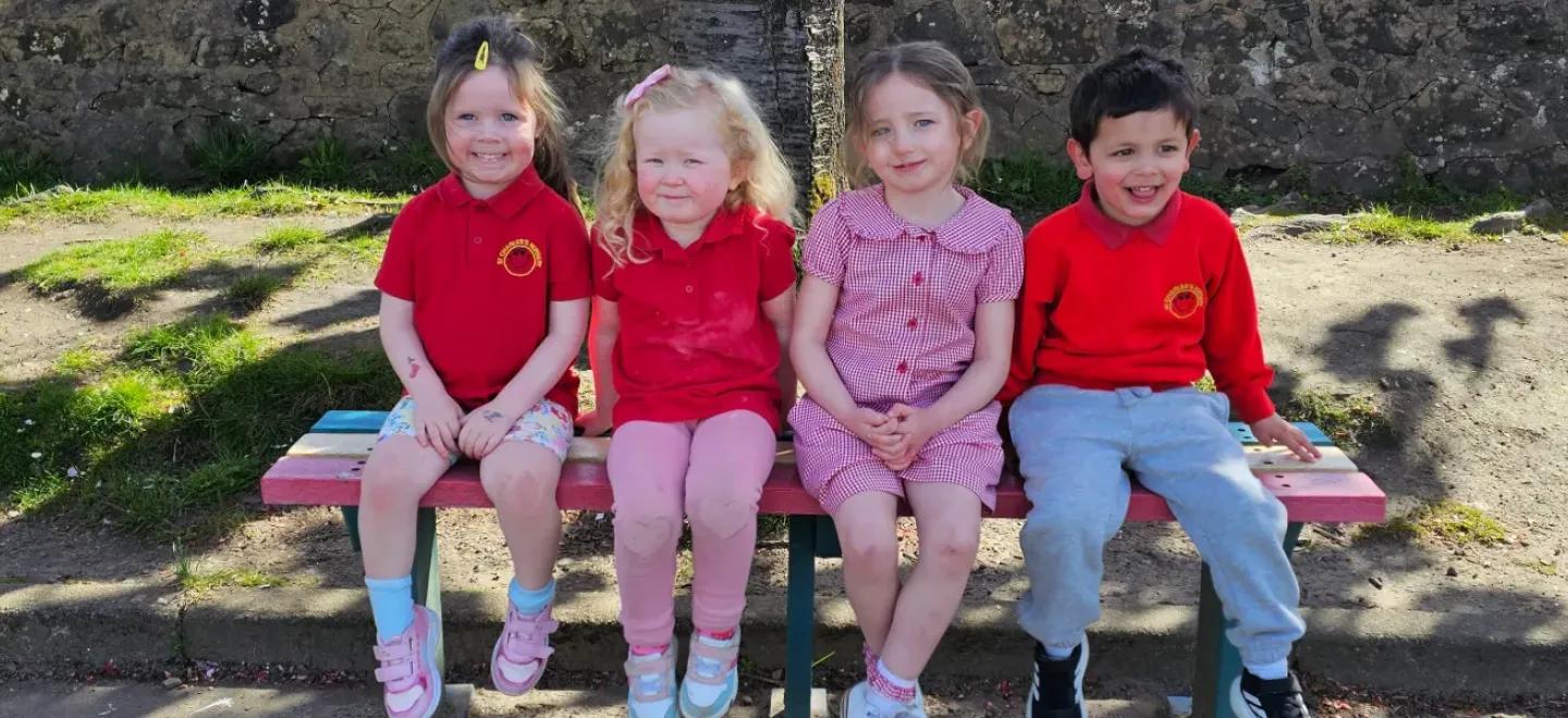 Four young children dressed in school uniform sit on a bench in their playground.