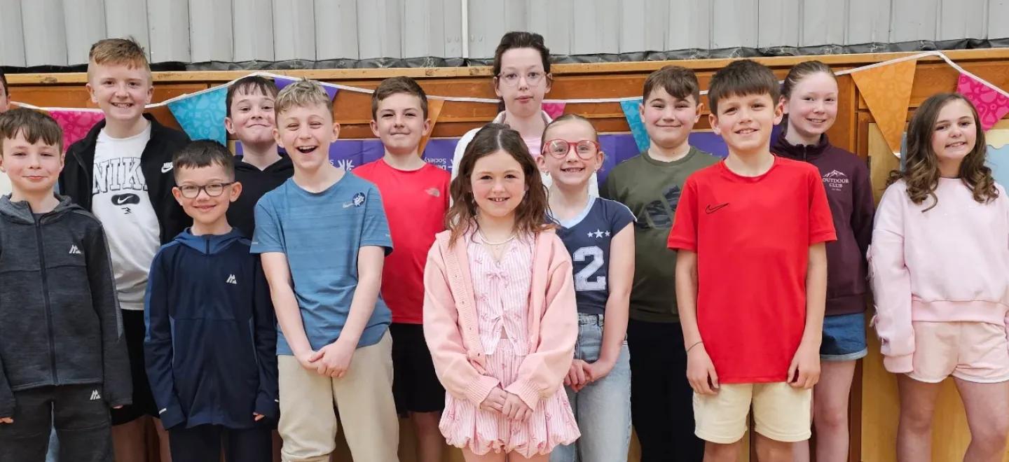 A group of senior primary school children smile while standing together in a hall.