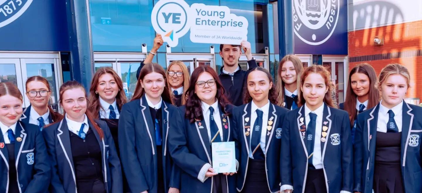 A large group of young people wearing school uniform stand together. A young person in the centre holds an award trophy.
