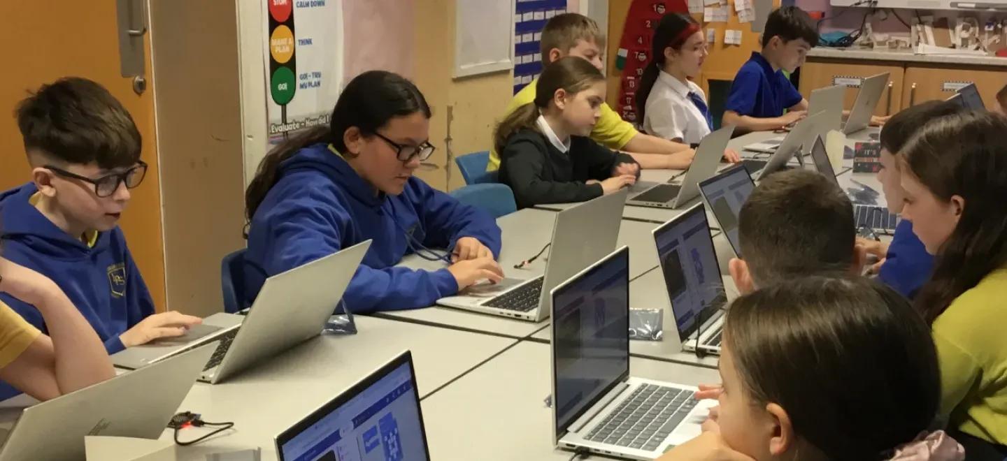 Pupils work on laptops seated around a large desk in a classroom.