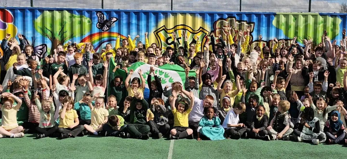 Whole school stand together in rows on grass pitch. Pupils in the middle hold a green banner.