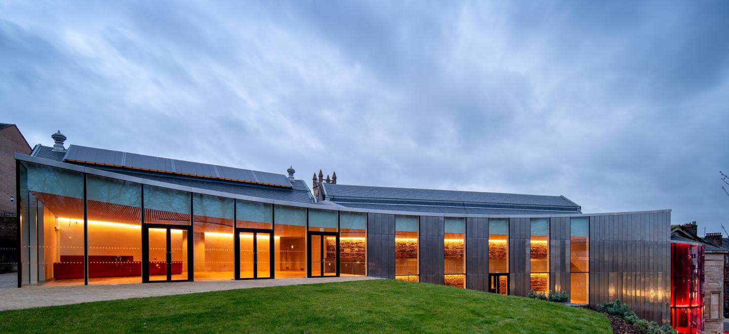 An image of the upper gallery from the garden of Paisley Museum. The glass windowed building is lit up in orange inside