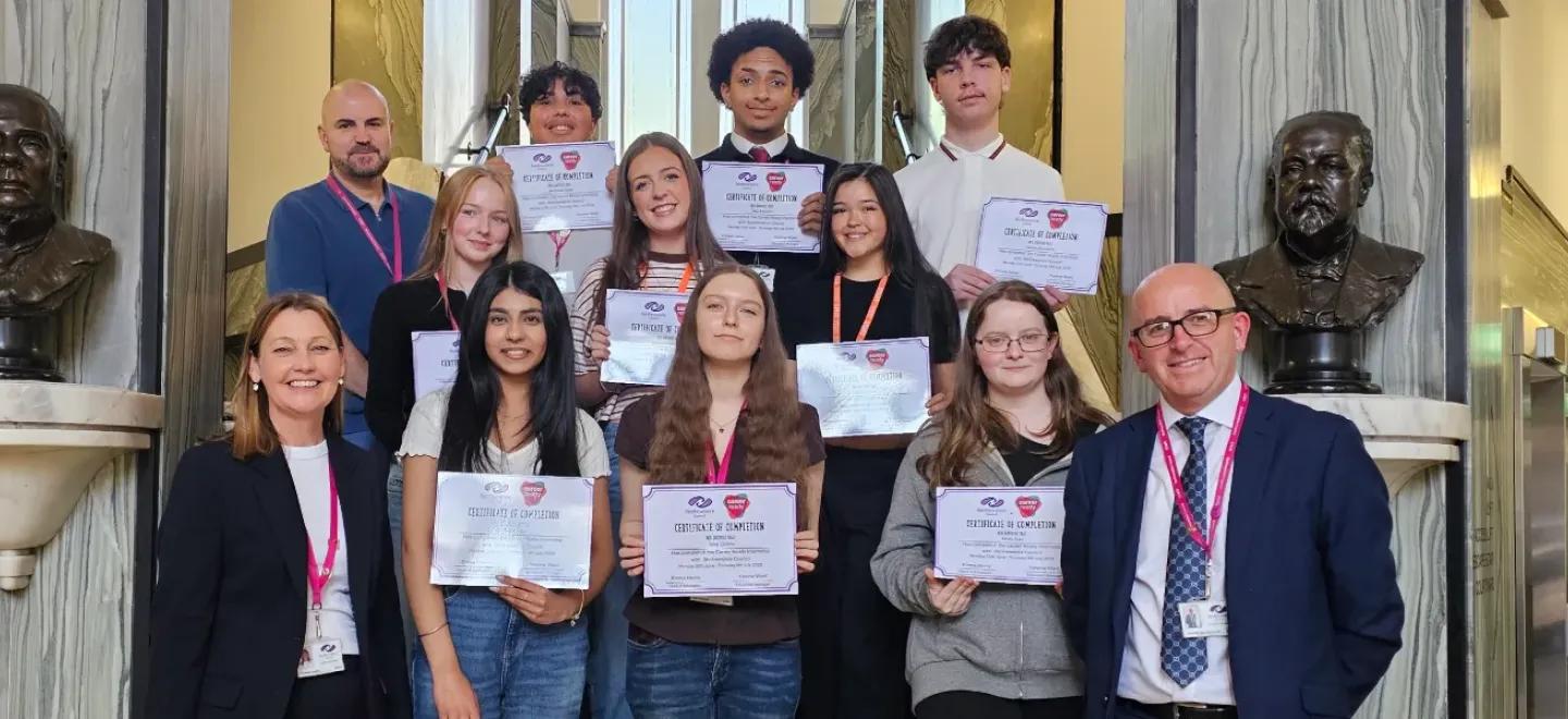 Young people hold certificates and stand alongside their mentors and Council representatives on stairs inside the Russell Institute building.